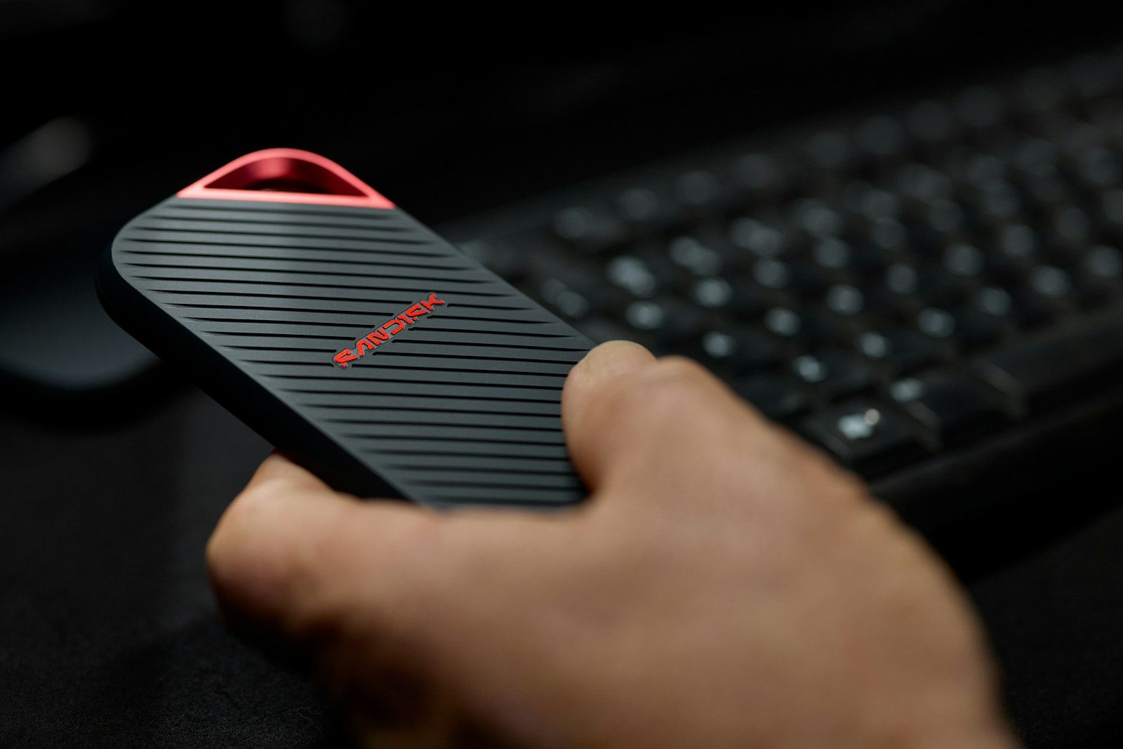 Hand holds black device with red light near keyboard.