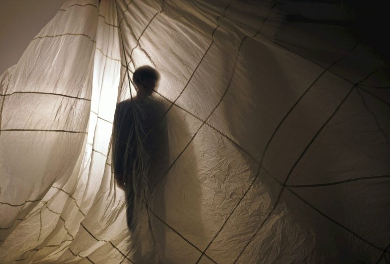 Person's dark silhouette visible through a backlit, grid-patterned white fabric.